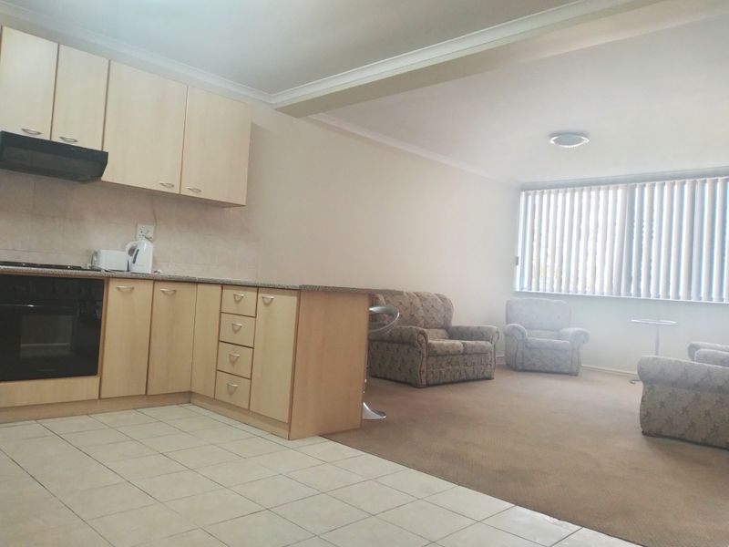 Open plan kitchen and living area featuring light wood cabinetry, granite countertops, built-in oven, tiled kitchen flooring, and carpeted living space with vertical blinds.