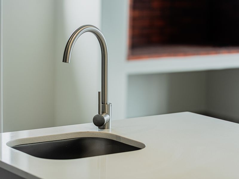 A close-up view of a modern kitchen island featuring a sleek stone countertop, undermount sink, and contemporary mixer tap with a fireplace in the background.
