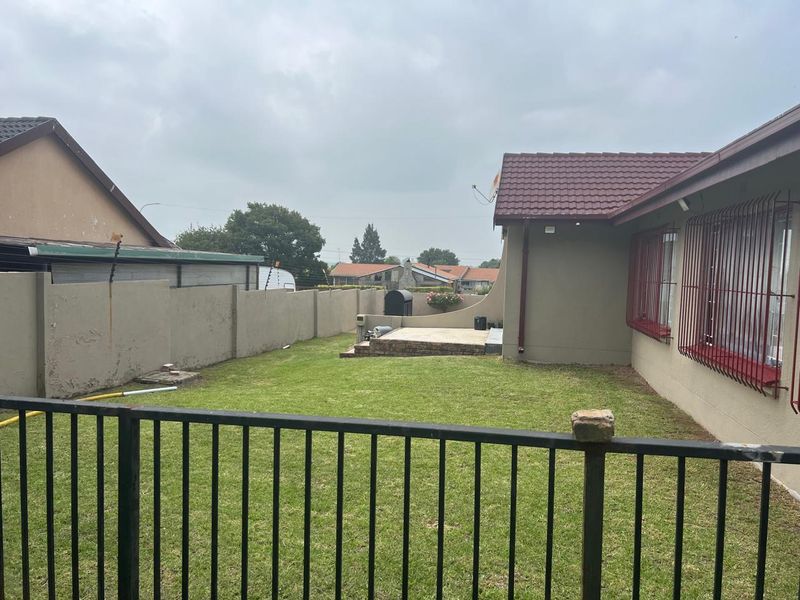 A rear garden view featuring a lawn, a small paved patio area, perimeter boundary walls, and security bars on the house windows.