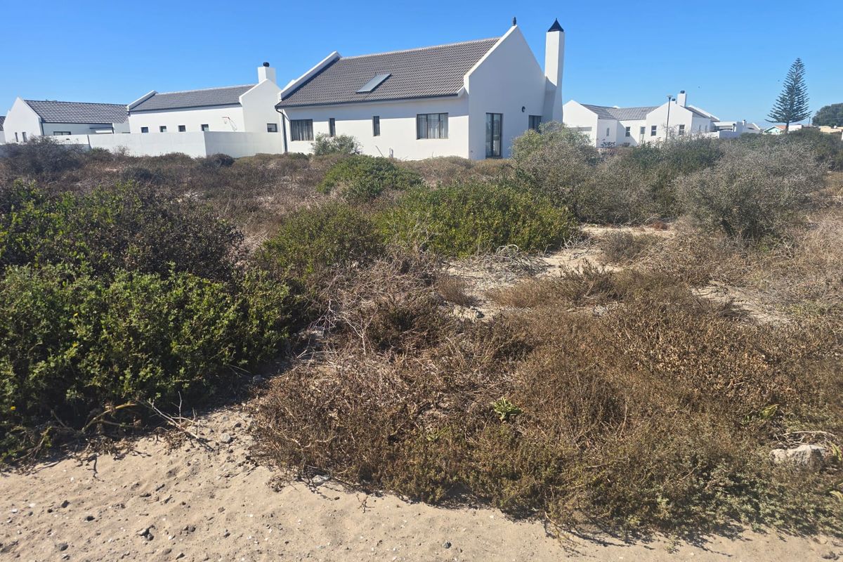 A view of a vacant residential plot featuring natural coastal vegetation, with established white-walled homes visible in the background.