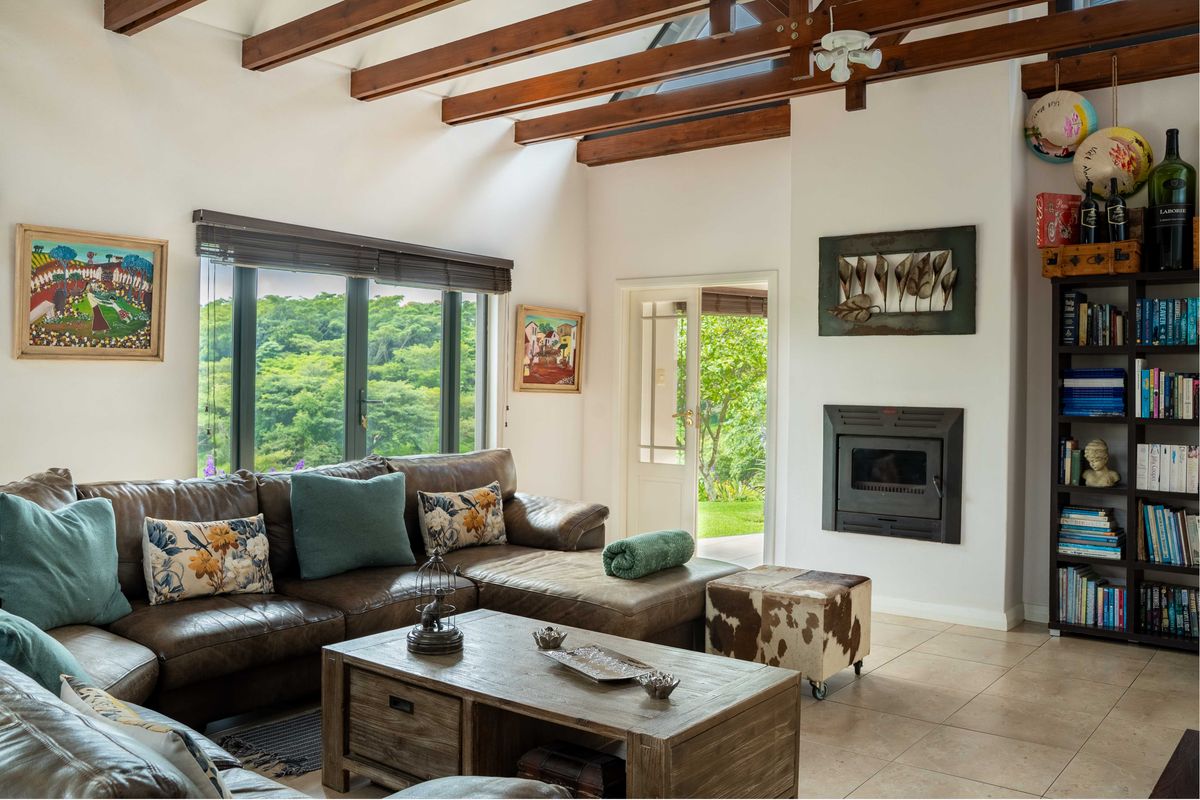 Cozy living room featuring exposed timber ceiling beams, a built-in fireplace, tiled flooring, and large windows providing natural light and garden views.