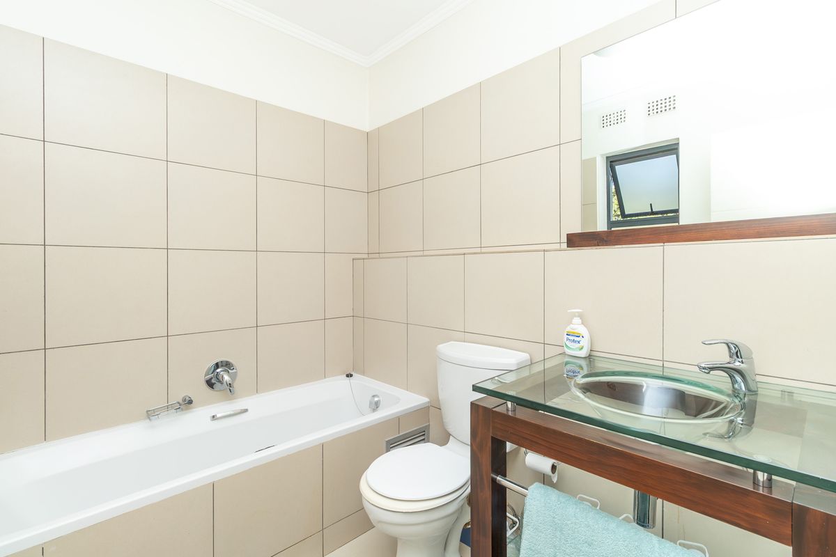 A functional bathroom featuring neutral wall tiling, a built-in bathtub, a toilet, and a modern glass-top vanity with a stainless steel basin.