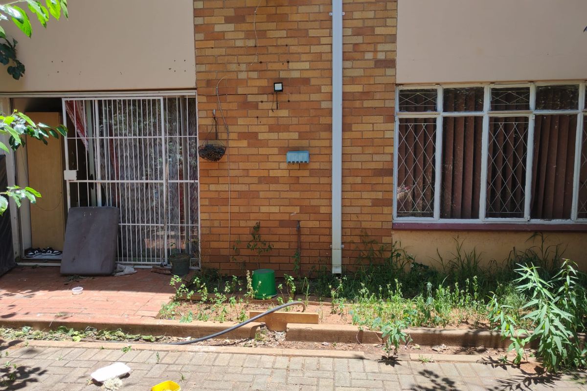 Rear exterior view of the apartment featuring a brick facade, paved patio area, and security-fitted windows and doors, requiring maintenance and landscaping.
