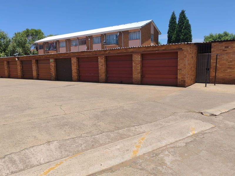 A row of brick-built garages with individual doors, situated in a paved parking area with a multi-story apartment building in the background.
