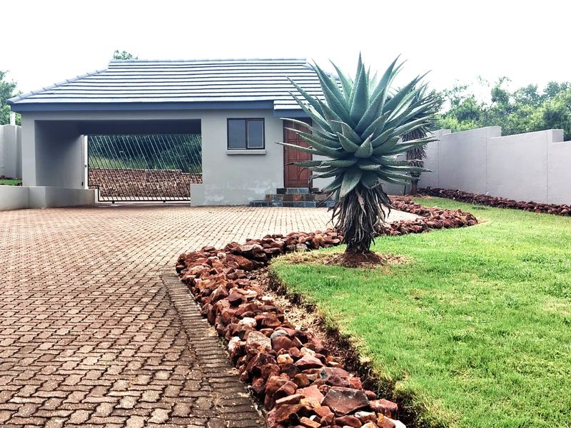 Front view of the property featuring a paved driveway, covered carport, landscaped garden with rock borders, and a secure perimeter wall.