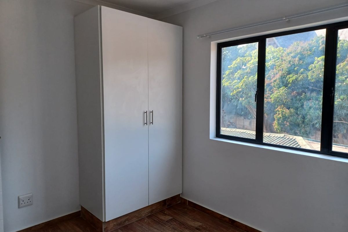 A bright, empty bedroom featuring modern laminate flooring, a built-in white wardrobe, and a large window with black aluminium frames.