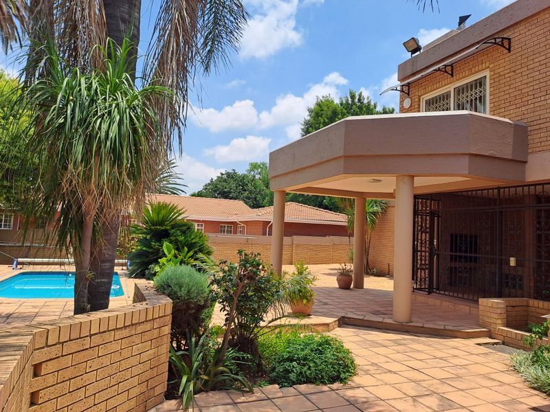 Rear exterior view featuring a covered patio area, brick paving, and a swimming pool set within a landscaped garden.