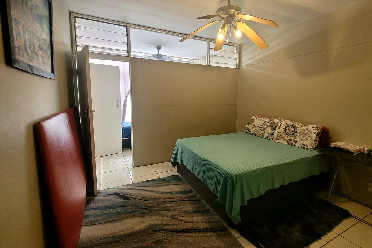 A functional bedroom featuring tiled flooring, a ceiling fan, and louvre windows providing natural light and ventilation.