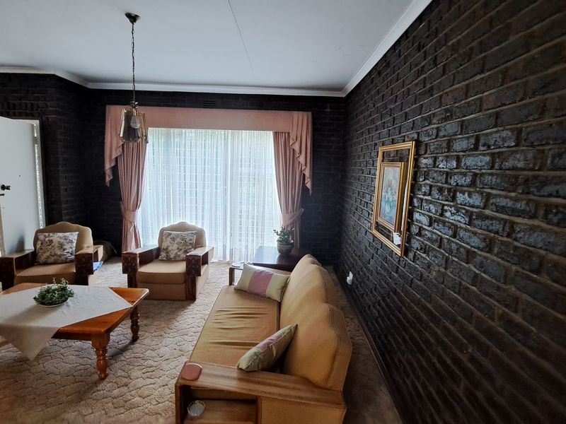 A living room featuring dark exposed brick walls, neutral carpeting, and a large window with draped curtains, offering a classic, dated aesthetic.