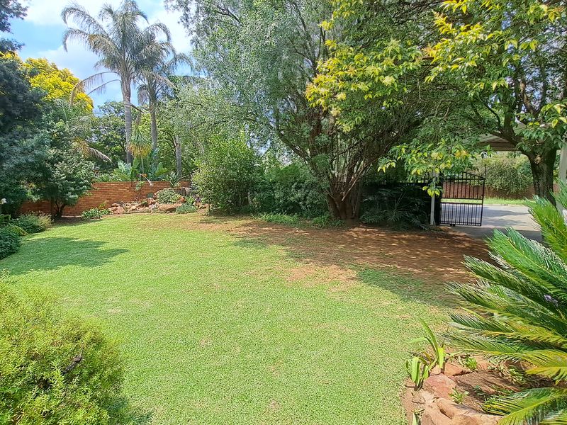 A well-maintained, spacious garden featuring mature trees, a manicured lawn, rockery landscaping, and a secure metal gate entrance.