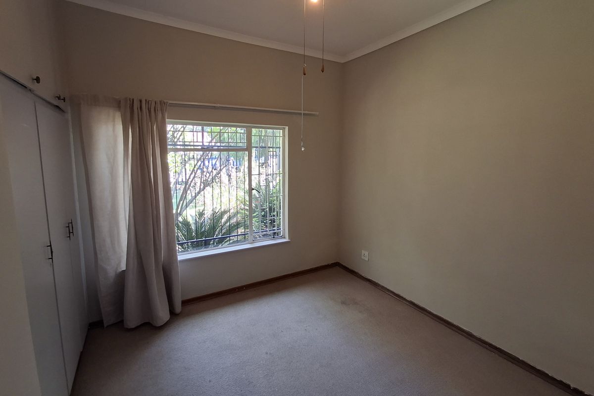 An empty bedroom featuring built-in cupboards, a ceiling fan, and security bars on the window. The room requires cosmetic updates to the flooring and paint.