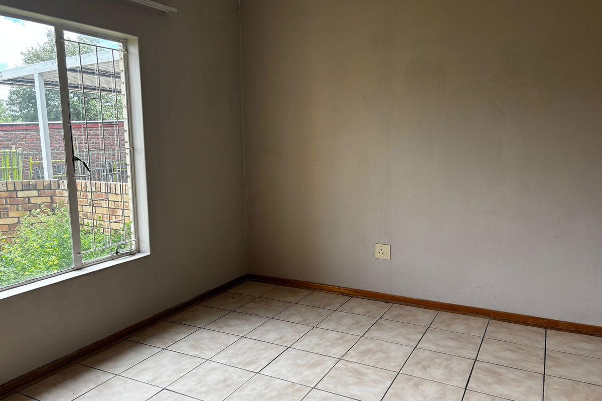 An empty bedroom featuring neutral tiled flooring, a large window with security bars, and a standard electrical outlet.