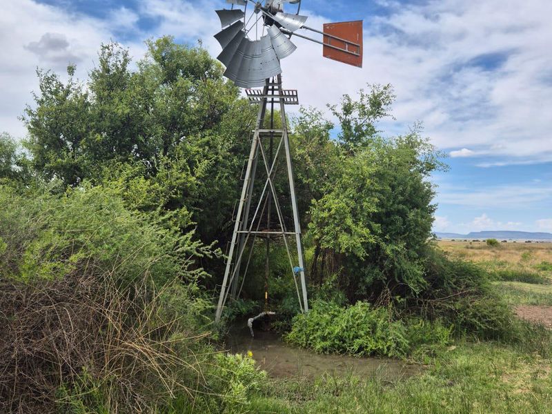 A traditional metal farm windmill standing in a rural landscape, featuring a functional water pumping mechanism and surrounding natural vegetation.