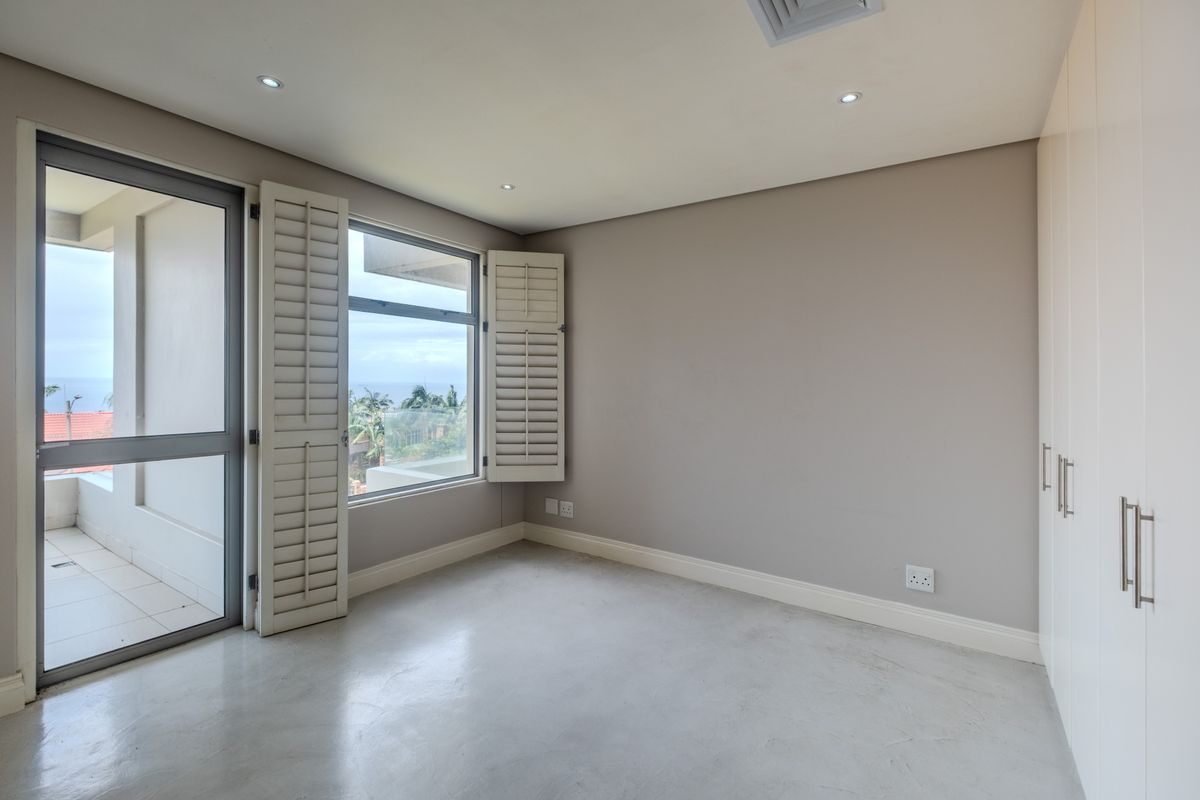 A bright, modern bedroom featuring polished concrete floors, built-in cupboards, plantation shutters, and direct access to a private balcony with scenic views.