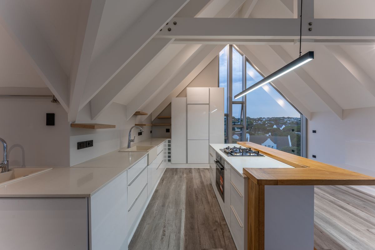 Modern attic kitchen featuring vaulted ceilings, sleek white cabinetry, stone countertops, a central island with a gas hob, and a timber breakfast bar.