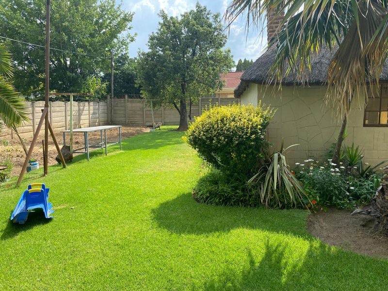 A well-maintained garden area featuring a lawn, established shrubs, and a structure with a traditional thatched roof, enclosed by a precast concrete boundary wall.