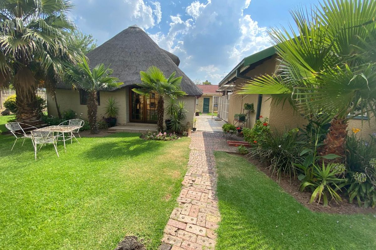 A well-maintained rear garden featuring a thatched roof structure, a brick paved pathway, and established landscaping.
