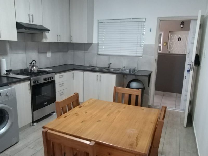 A functional kitchen featuring light-toned cabinetry, a gas hob with extractor fan, double sink, and tiled backsplash, with direct access to an adjacent hallway.