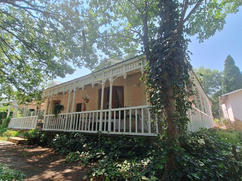 A charming historic house featuring a wrap-around veranda with decorative timber fretwork, surrounded by mature trees and established garden beds.