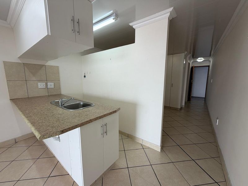 A functional kitchen area featuring laminate countertops, stainless steel sink, and tiled flooring, leading into a hallway with built-in storage cupboards.