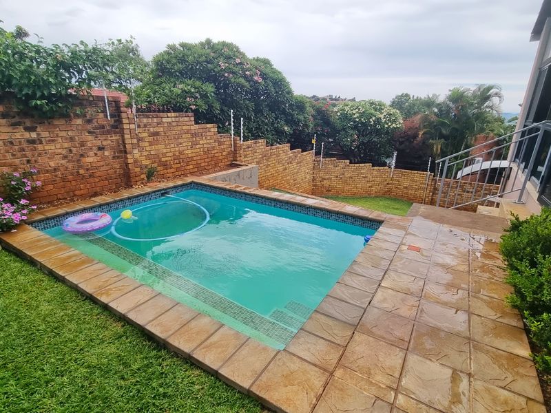 A compact swimming pool area featuring stone-tiled coping, a surrounding lawn, and a brick boundary wall with integrated security fencing.