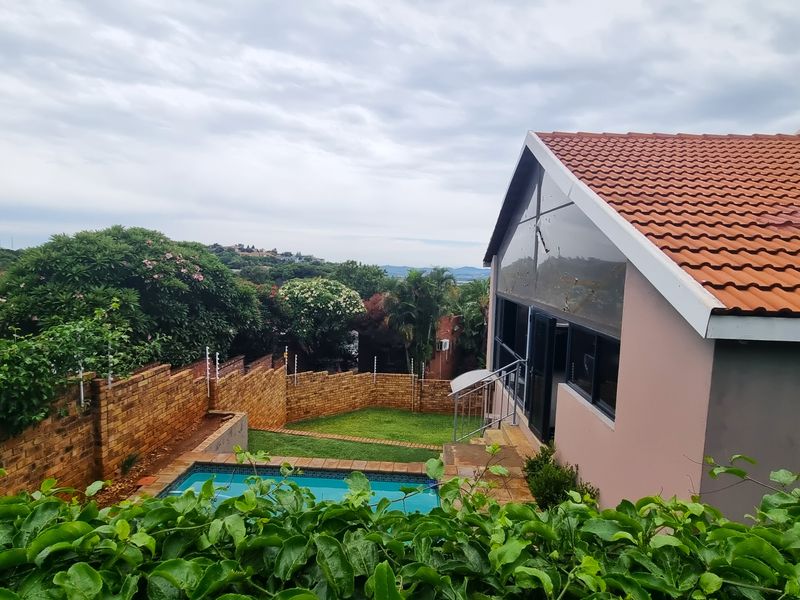 Rear view of the property featuring a swimming pool, landscaped garden, brick boundary wall, and a pitched roof structure with large glass windows.