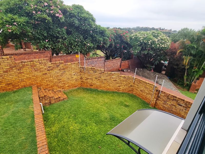 A private rear garden featuring tiered brick retaining walls, established trees, and a small covered patio area.