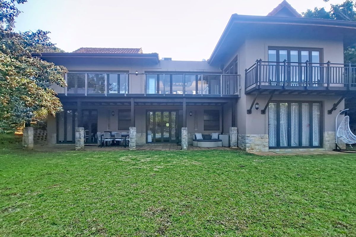Rear view of a two-story house featuring a spacious lawn, ground-floor patio, and upper-level balcony with glass balustrades and stone facade accents.