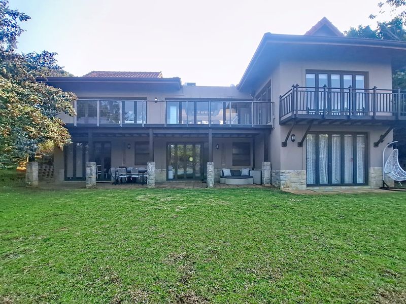 Rear view of a two-story house featuring a spacious lawn, ground-floor patio, and upper-level balcony with glass balustrades and stone facade accents.