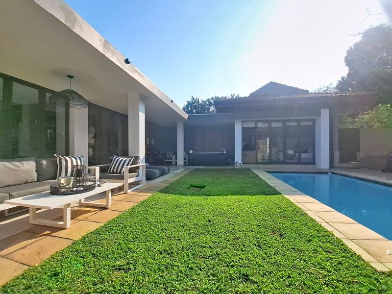 A well-maintained outdoor entertainment area featuring a swimming pool, a covered patio with paved flooring, and a manicured lawn.