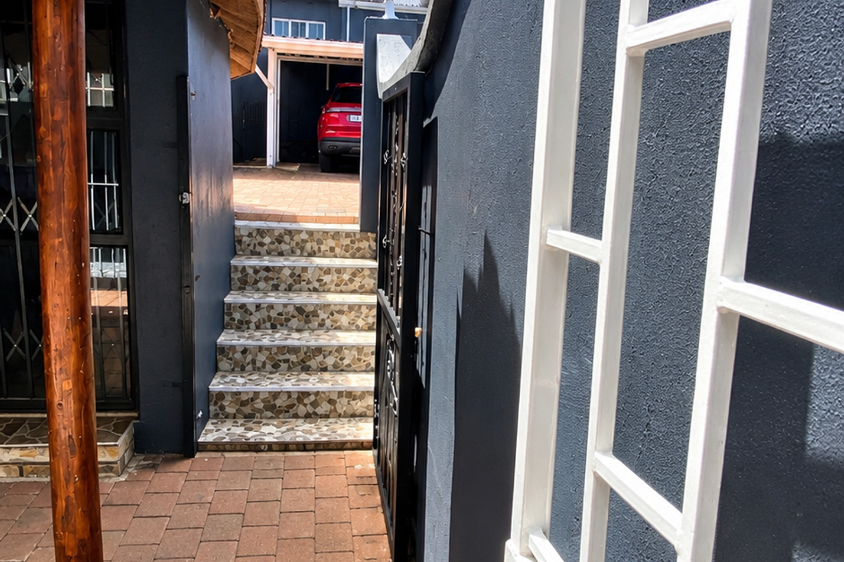 A paved outdoor walkway featuring a thatched roof structure, tiled stairs, and a secure metal gate leading to a rear parking area.