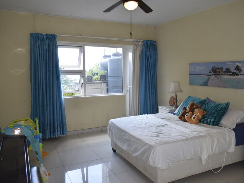 A spacious bedroom featuring tiled flooring, a ceiling fan, and large windows providing natural light and access to an outdoor area.