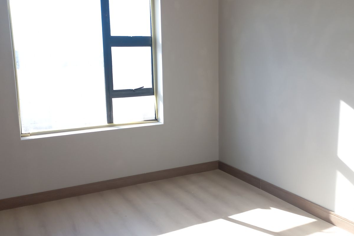 A bright, empty bedroom featuring neutral walls, laminate flooring, and a large aluminium-framed window allowing for ample natural light.