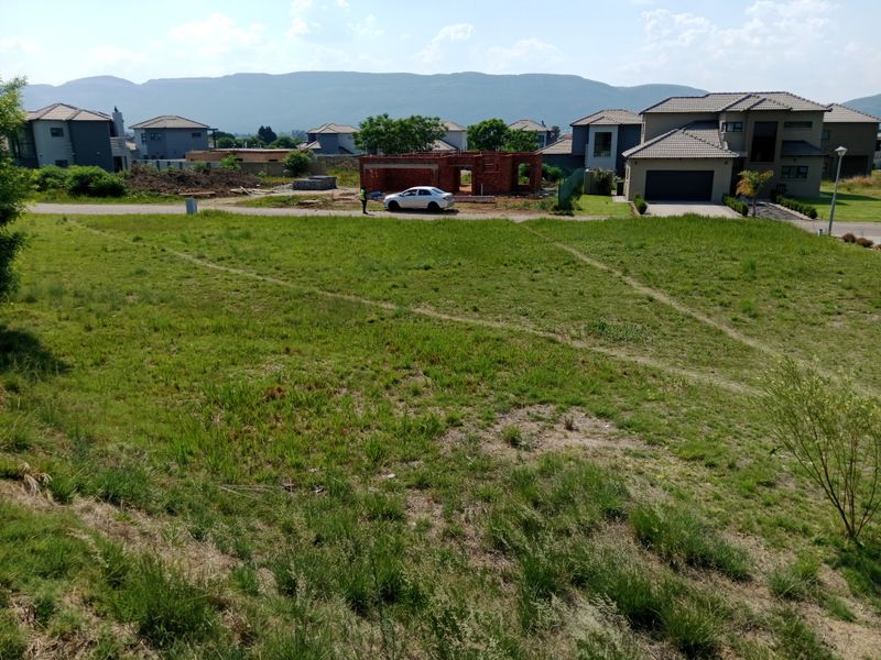 A vacant residential plot of land featuring natural grass cover, situated in a developed estate with surrounding residential homes and mountain views.