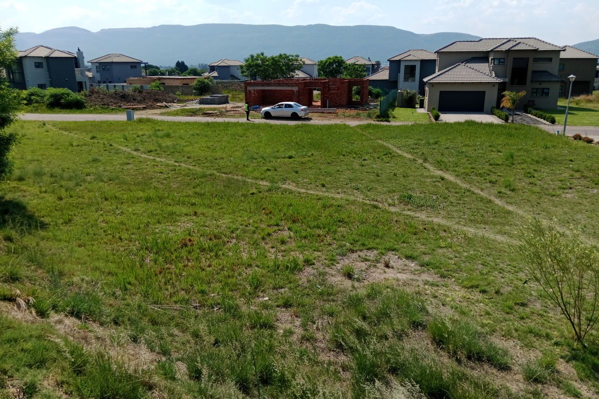 A vacant residential plot of land featuring natural grass cover, situated in a developed estate with surrounding residential homes and mountain views.