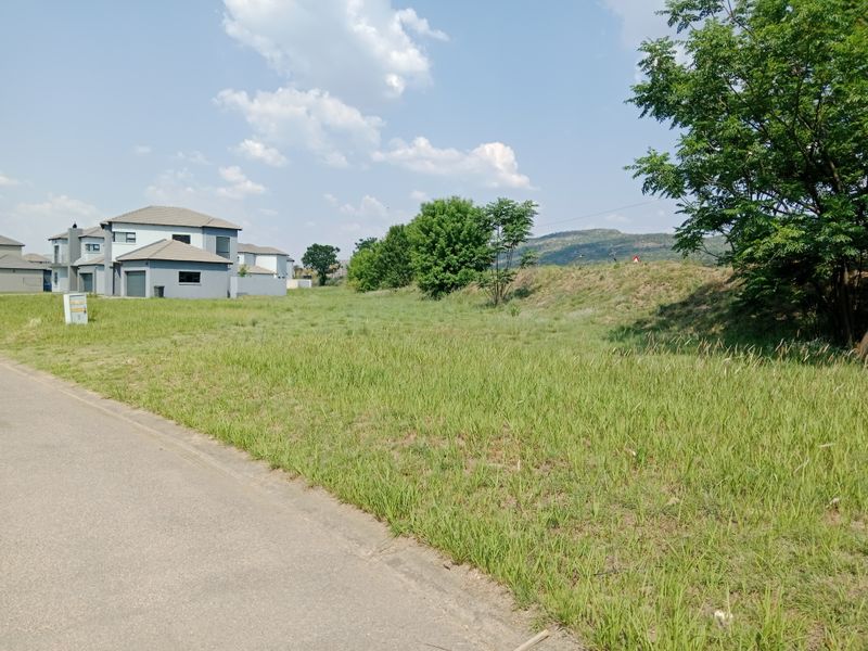 A view of a vacant residential plot from the adjacent paved street, featuring a grassy slope and established trees in a suburban estate setting.