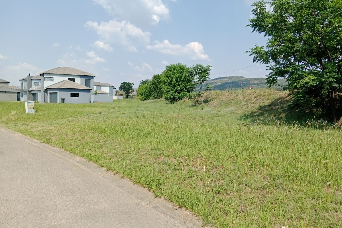 A view of a vacant residential plot from the adjacent paved street, featuring a grassy slope and established trees in a suburban estate setting.