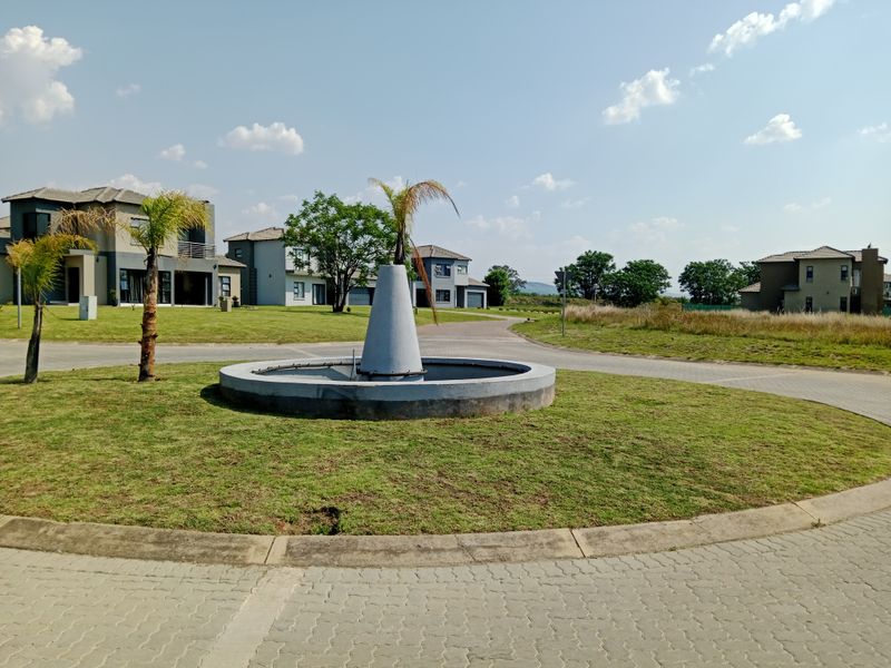 A view of a residential estate entrance featuring a central concrete fountain, paved driveway, and well-maintained landscaped lawn area.