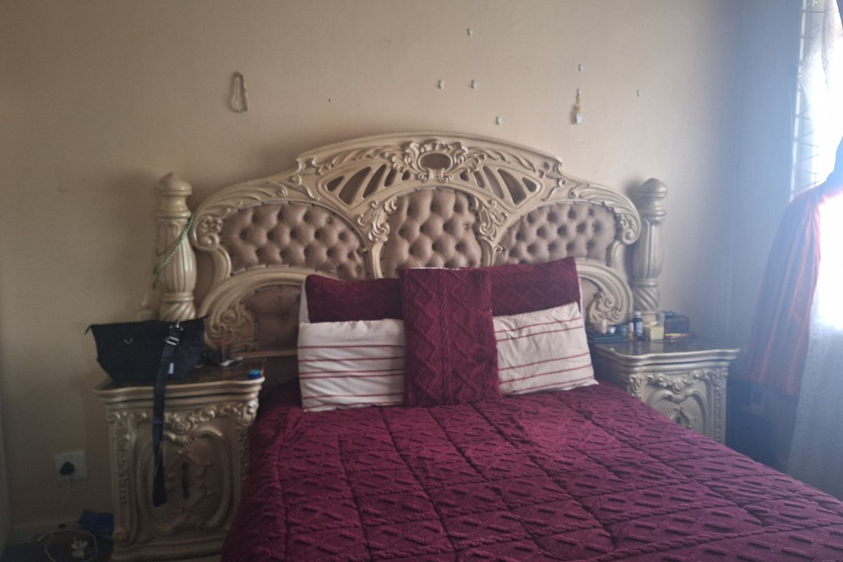 A bedroom featuring a large ornate headboard and matching bedside tables, with visible wall surface imperfections requiring maintenance.
