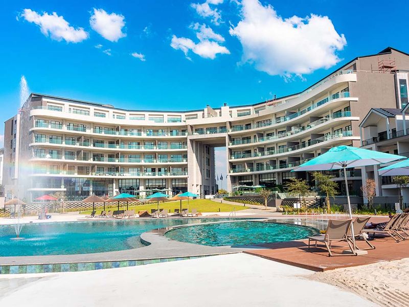 A well-maintained apartment complex featuring a large swimming pool with a water fountain, wooden decking, and landscaped outdoor areas.