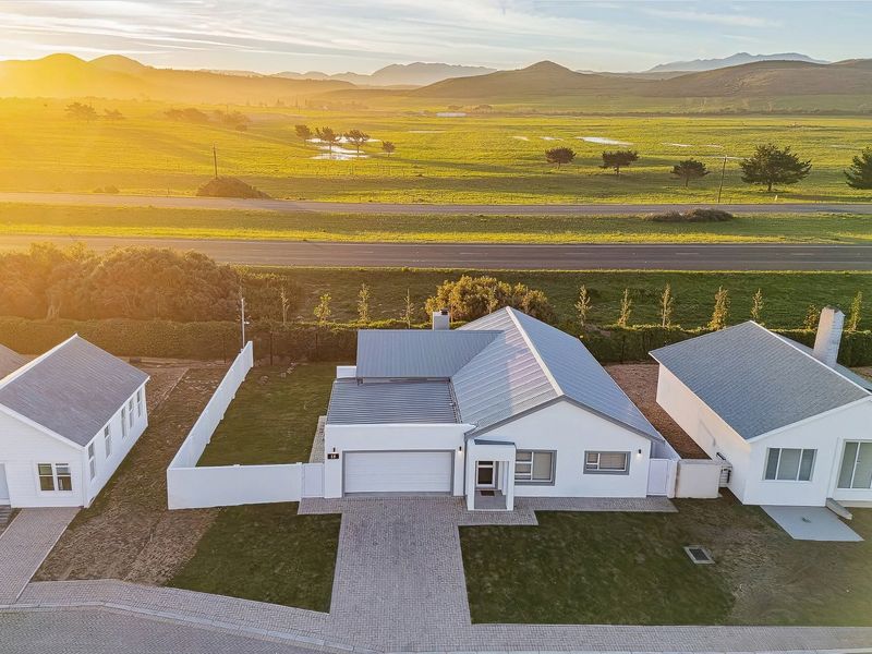 Aerial view of a modern single-story house with a metal roof, paved driveway, and surrounding boundary walls, set against a scenic rural landscape.
