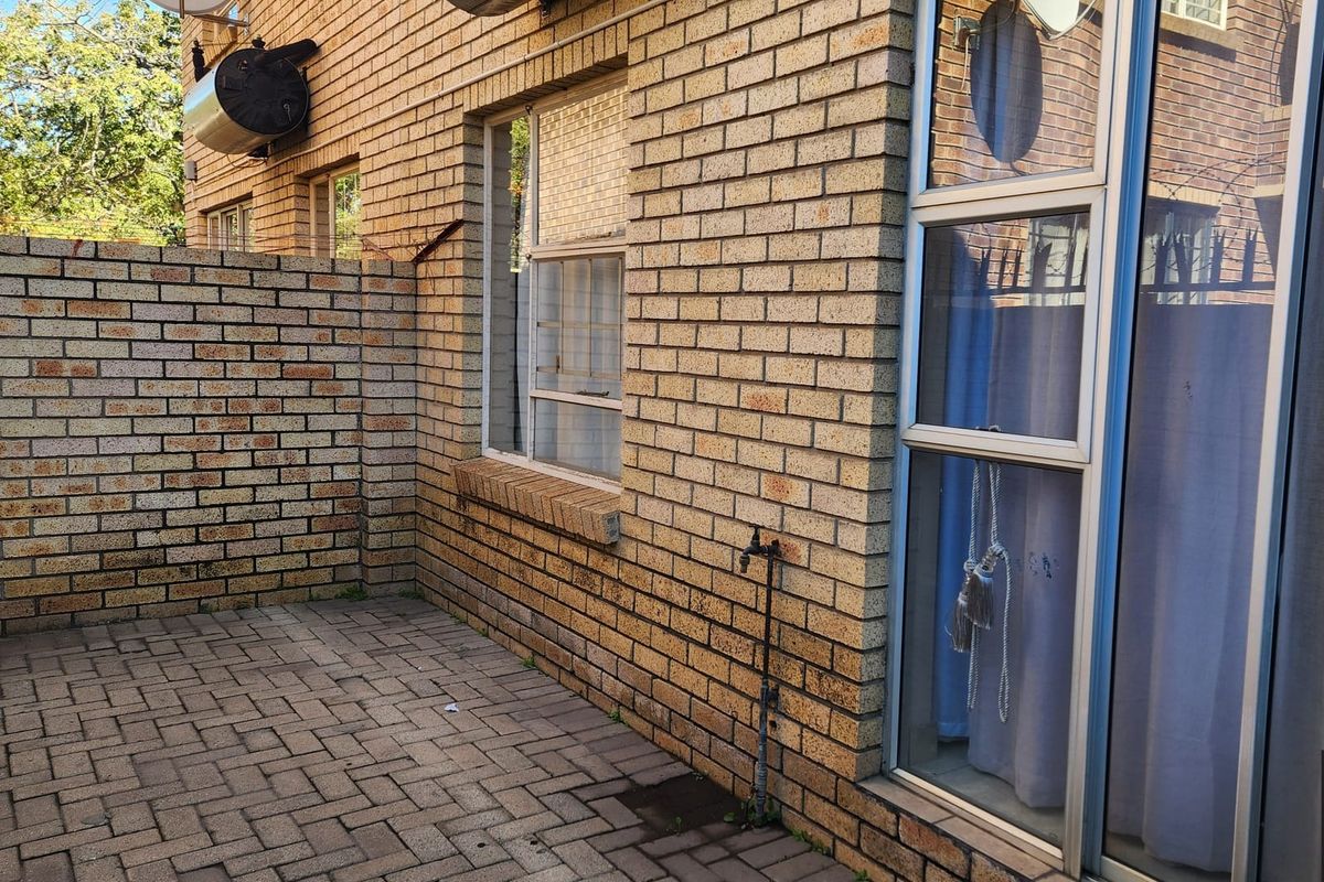 A rear exterior view of the apartment featuring a paved patio area, brick facade, external water tap, and wall-mounted geyser.