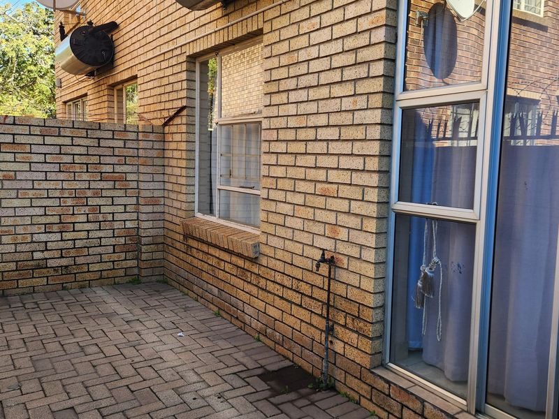 A rear exterior view of the apartment featuring a paved patio area, brick facade, external water tap, and wall-mounted geyser.