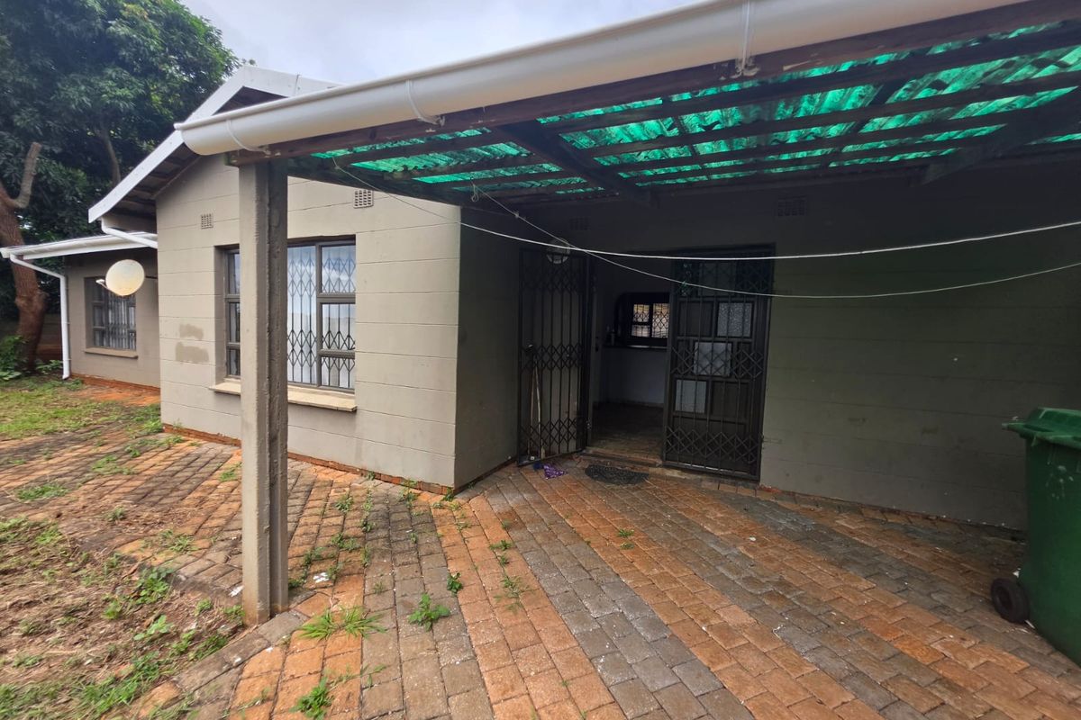 Rear exterior view featuring a covered patio area with brick paving and security gates, showing signs of wear and maintenance requirements.