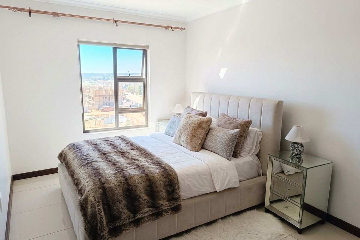 A bright and well-maintained bedroom featuring neutral tiled flooring, a large aluminium-framed window, and coved ceiling detailing.