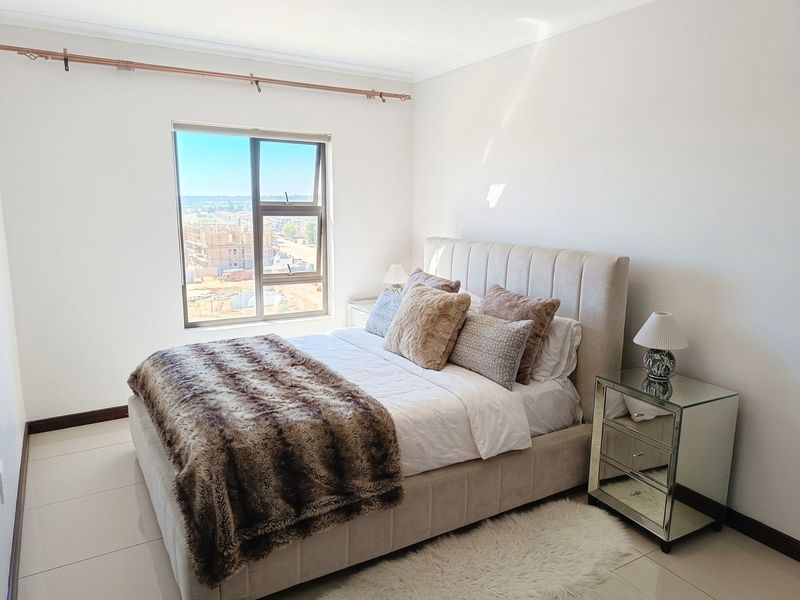 A bright and well-maintained bedroom featuring neutral tiled flooring, a large aluminium-framed window, and coved ceiling detailing.