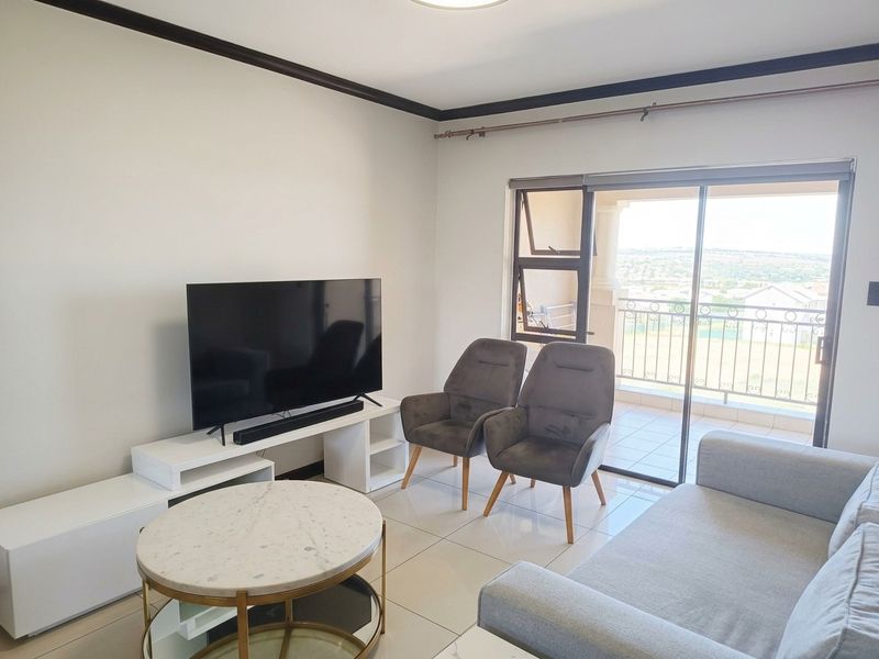 A bright living room featuring tiled flooring, crown molding, and a sliding glass door providing direct access to a balcony with scenic views.