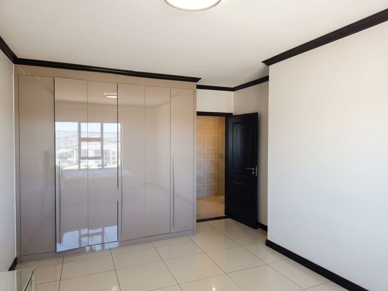 A bright, modern bedroom featuring high-gloss built-in cupboards, neutral tiled flooring, and dark contrasting trim, with direct access to an en-suite bathroom.
