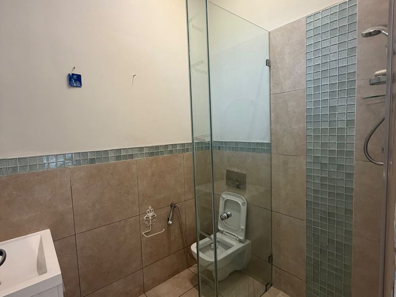 A functional bathroom featuring a glass-enclosed shower, wall-mounted toilet, and a vanity basin with neutral ceramic tiling and mosaic accents.