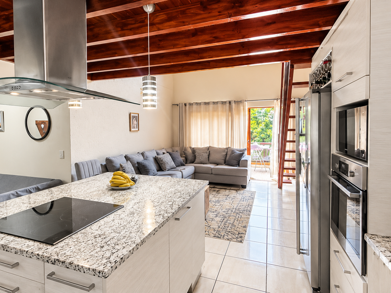 Spacious open-plan living area featuring a modern kitchen with granite countertops, built-in appliances, and striking exposed timber ceiling beams leading to a balcony.